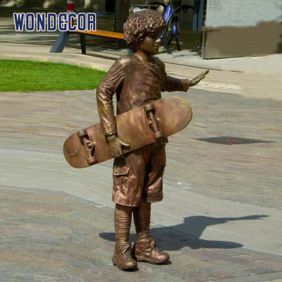 A bronze metal art sculpture of a boy holding a skateboard outdoors
