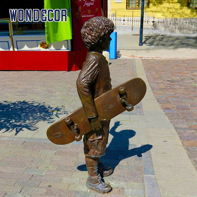 A bronze metal art sculpture of a boy holding a skateboard outdoors
