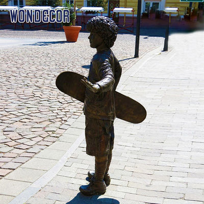 A bronze metal art sculpture of a boy holding a skateboard outdoors
